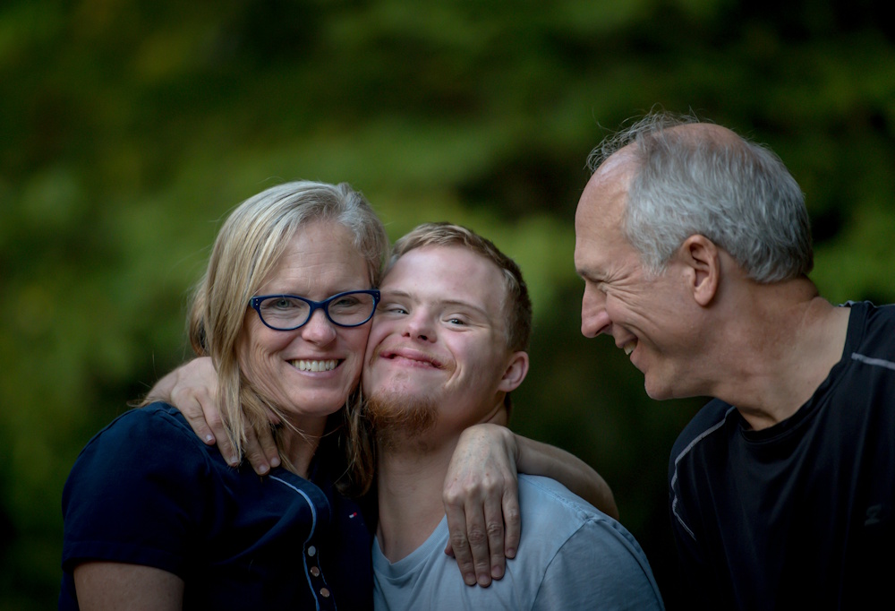 A smiling family of three, with a woman in glasses and a young man with Down syndrome looking at the camera, and the father looking fondly at them, all against a blurred green background.