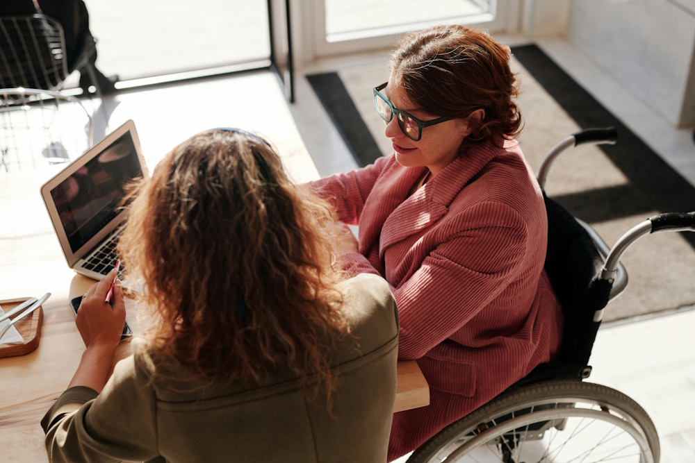 a woman sitting with another woman in a wheelchair in front of a laptop